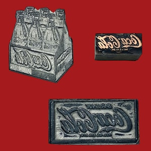May include: Three vintage Coca-Cola printing blocks. One block depicts a six-pack of bottles, another shows the Coca-Cola logo, and the third block displays the Coca-Cola logo in reverse. All blocks are gray.