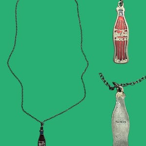 May include: A silver-toned necklace with a Coca-Cola bottle pendant. The pendant is red and white with the Coca-Cola logo. The back of the pendant is stamped with "TAIWAN". The necklace is on a green background.