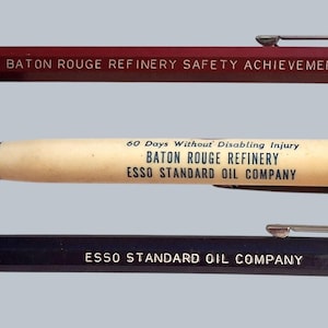 May include: Three vintage pencils with metal clips. One is red with "BATON ROUGE REFINERY SAFETY ACHIEVEMENT" printed on it. Another is cream-colored with "60 Days Without Disabling Injury BATON ROUGE REFINERY ESSO STANDARD OIL COMPANY". The last is navy blue with "ESSO STANDARD OIL COMPANY".