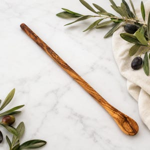 May include: A long, slender wooden stirring spoon crafted from olive wood, featuring a distinct natural grain pattern with swirling light and dark brown tones, a smooth finish, and a shallow, elongated oval bowl at the end of the handle.