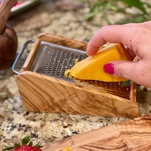 Peut inclure: Une râpe à fromage en bois avec un insert en acier inoxydable. Une main tient un morceau de fromage jaune au-dessus de la râpe.
