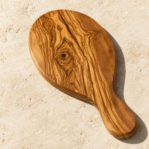 May include: A wooden serving board with a rounded top and a short handle, featuring a prominent natural wood grain pattern with swirling dark brown and light tan streaks and a distinct knot in the center of the surface.