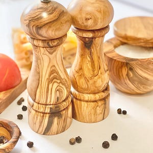 May include: A set of two wooden salt and pepper shakers with a natural wood grain finish. The shakers are made of olive wood and have a classic design.