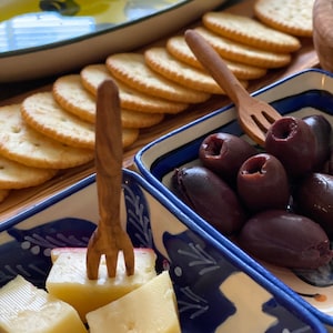 May include: A blue and white ceramic dish filled with olives, a blue and white ceramic dish filled with cubed cheese, and a wooden fork.  A wooden serving tray with a blue and white ceramic dish filled with crackers.