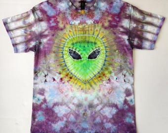 May include: A tie-dye t-shirt with a green alien face design in the center. The shirt has a purple, blue, and yellow color scheme.