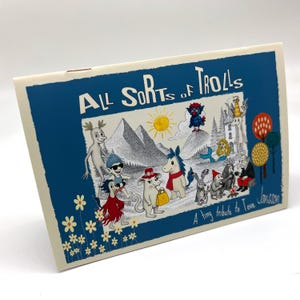 以下が含まれることがあります： 青い縁取りと白い文字で「All Sorts of Trolls」と書かれた長方形のカード。カードには、さまざまなトロルのような生き物、山、太陽のイラストが描かれています。「A tiny tribute to Tove Jansson」というテキストも表示されています。