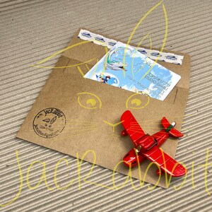 Peut inclure: Un petit avion rouge jouet est pos&eacute; sur une enveloppe en papier brun. L'enveloppe contient une carte Studio Ghibli et est scell&eacute;e avec du ruban adh&eacute;sif d&eacute;coratif. Un logo Jackrabbit est estampill&eacute; sur l'enveloppe.