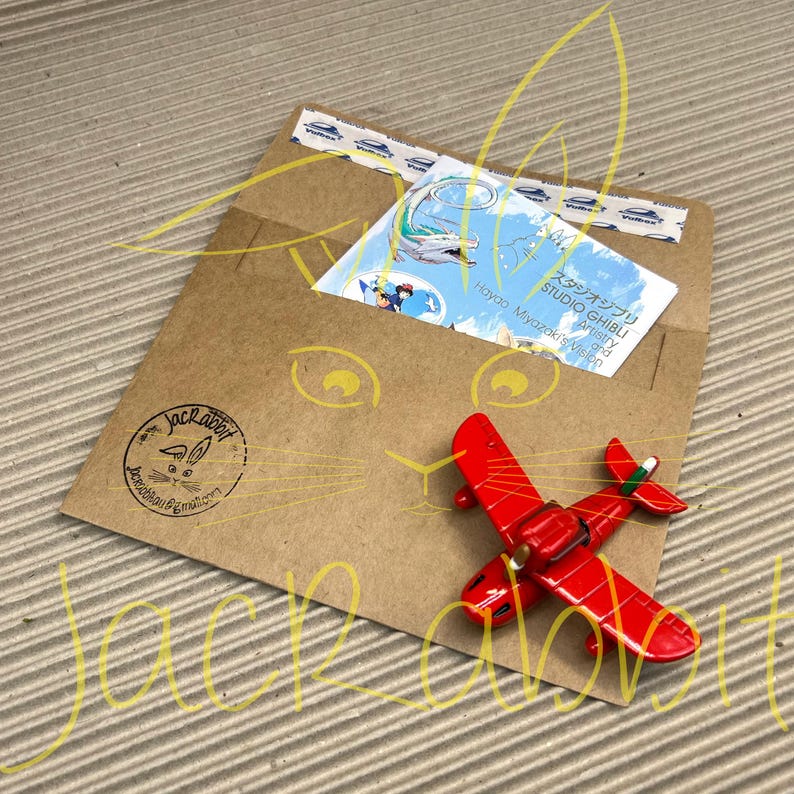 Peut inclure: Un petit avion rouge jouet est pos&eacute; sur une enveloppe en papier brun. L'enveloppe contient une carte Studio Ghibli et est scell&eacute;e avec du ruban adh&eacute;sif d&eacute;coratif. Un logo Jackrabbit est estampill&eacute; sur l'enveloppe.