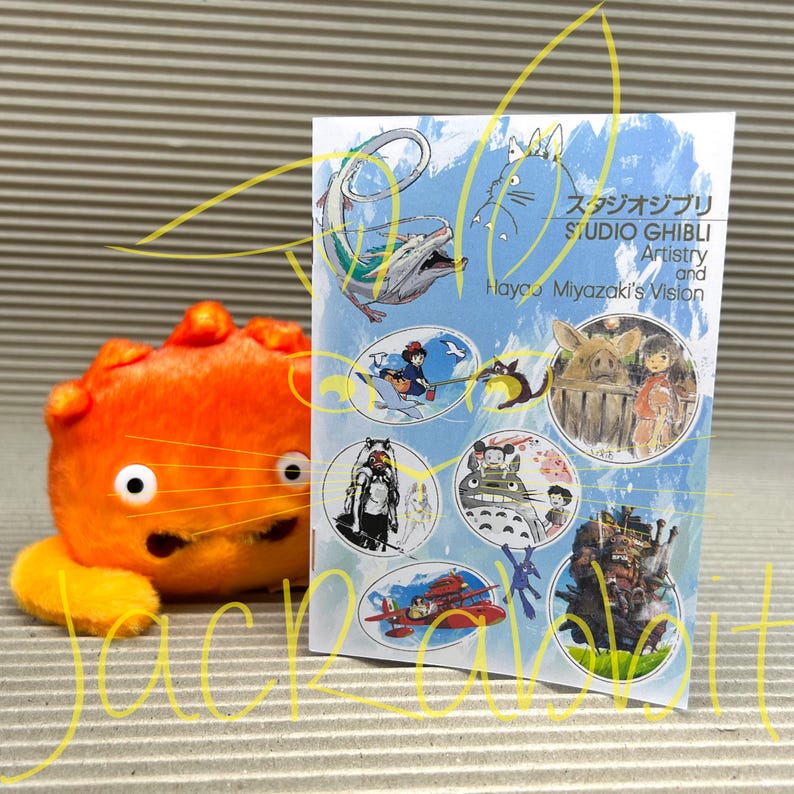 Peut inclure: Un jouet en peluche orange avec un corps rond et un visage souriant est &agrave; c&ocirc;t&eacute; d'un livret. Le livret pr&eacute;sente des illustrations de personnages de films du Studio Ghibli, avec le titre "STUDIO GHIBLI Artistry and Hayao Miyazaki's Vision."