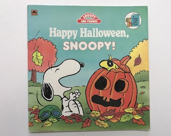 May include: A children's book cover featuring Snoopy and friends celebrating Halloween. The cover shows Snoopy peeking out from behind a large orange pumpkin with a jack-o'-lantern face. The text on the cover reads "Happy Halloween, Snoopy!"