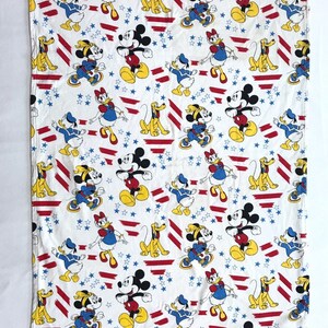 May include: White fabric with a repeating pattern of Mickey Mouse, Minnie Mouse, Donald Duck, and Pluto. The characters are wearing red, white, and blue outfits and are surrounded by red and white stripes and blue stars.