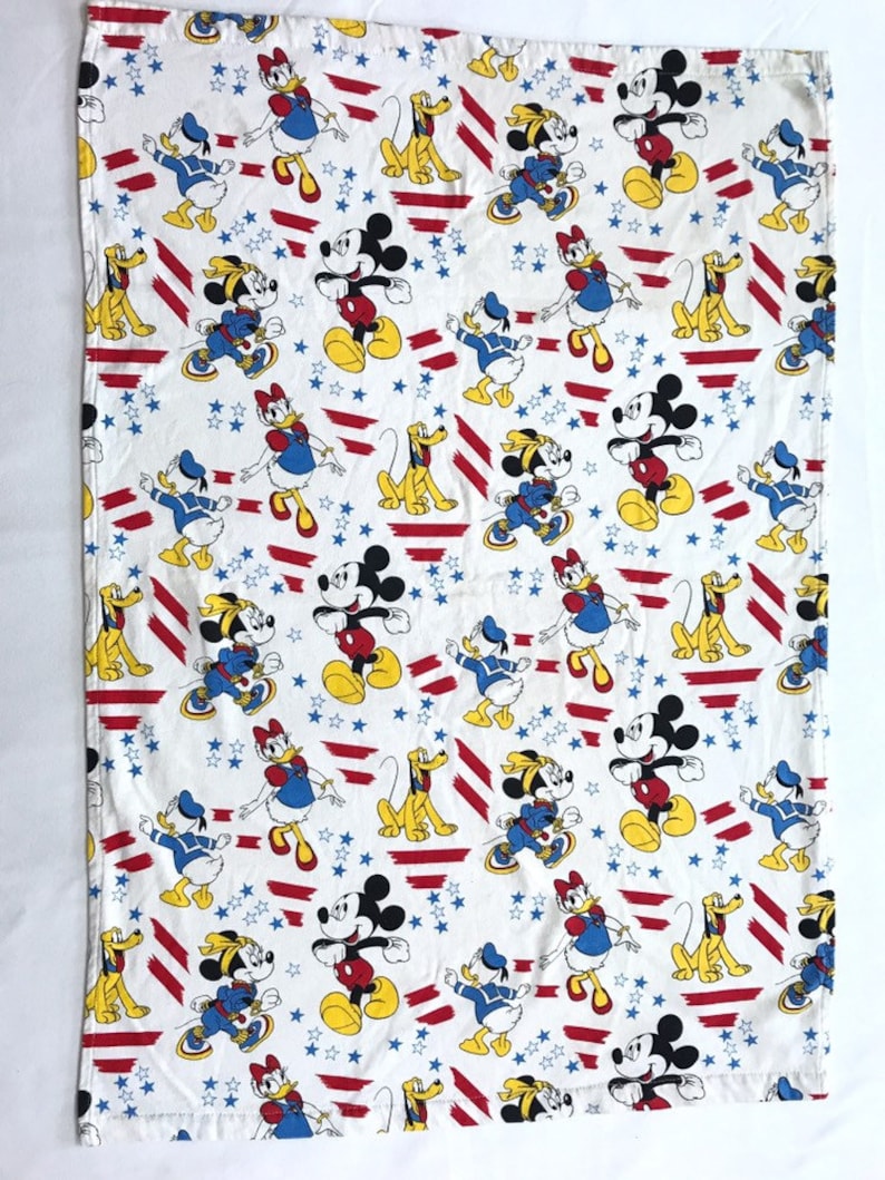 May include: White fabric with a repeating pattern of Mickey Mouse, Minnie Mouse, Donald Duck, and Pluto. The characters are wearing red, white, and blue outfits and are surrounded by red and white stripes and blue stars.
