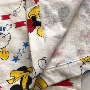May include: White fabric with a red, white and blue pattern featuring Mickey Mouse and Donald Duck. The fabric has a red, white and blue striped border.
