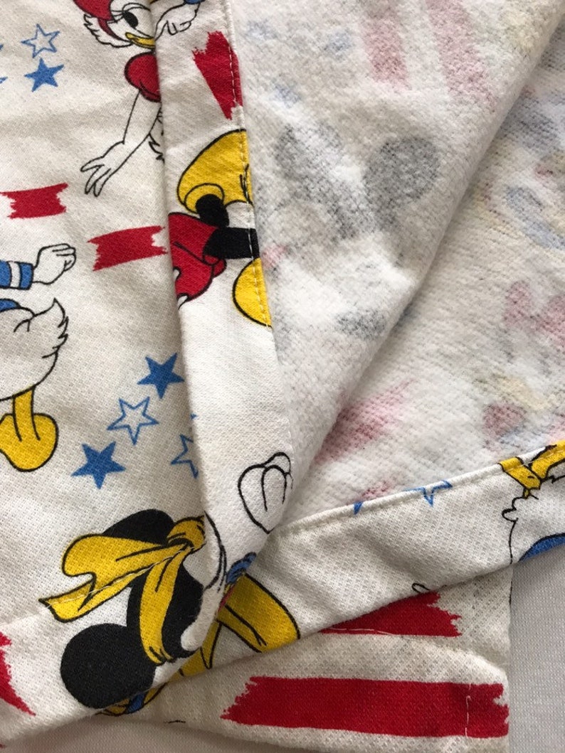 May include: White fabric with a red, white and blue pattern featuring Mickey Mouse and Donald Duck. The fabric has a red, white and blue striped border.