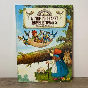 May include: A hardback children's book titled A Trip to Granny Rumbletummy's, illustrated by John Patience. The cover features a colourful illustration of a small figure in a hammock suspended by birds, and an elderly figure with a cane and two rabbits.