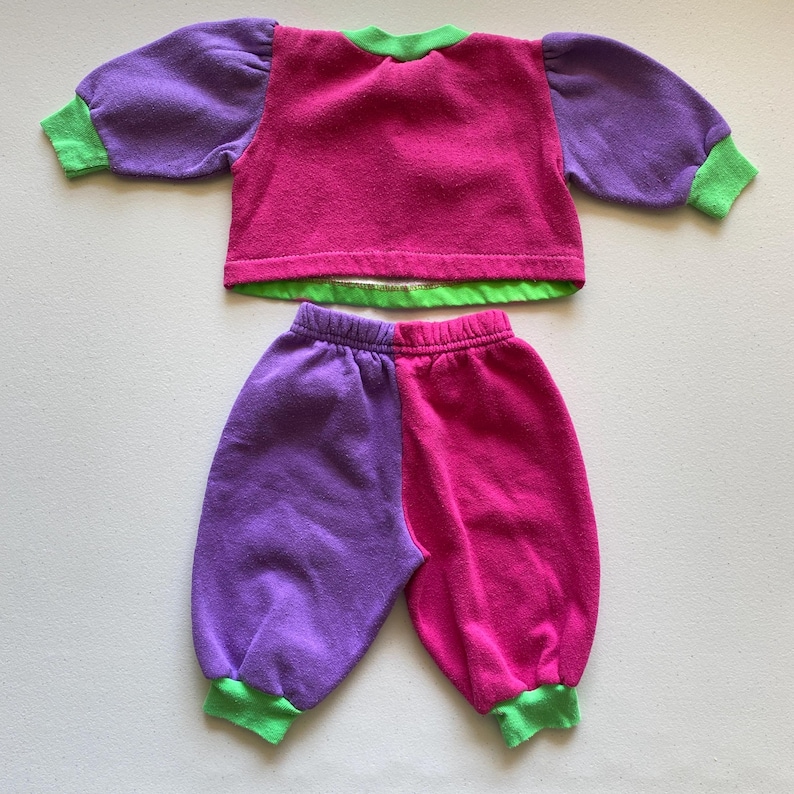 K&ouml;nnte beinhalten: Ein zweiteiliges Kinderoutfit in leuchtenden Farben. Das Oberteil ist magenta mit lila &Auml;rmeln und gr&uuml;nen Verzierungen. Die Hose ist halb lila und halb magenta, mit gr&uuml;nen B&uuml;ndchen. Das Outfit ist aus einem weichen, m&ouml;glicherweise fleeceartigen Material.