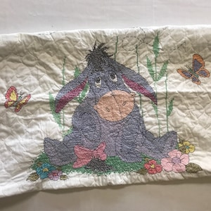 May include: A white quilted pillowcase with a cross-stitch design of a gray Eeyore from Winnie the Pooh. Eeyore is sitting in a field of green grass with pink, yellow, and orange flowers. There are two yellow butterflies on the pillowcase.