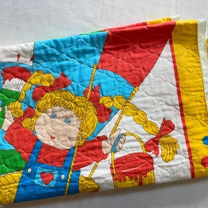 May include: A vintage, quilted baby blanket featuring a cartoon girl with blonde pigtails, red bows, and blue overalls. The design includes a paintbrush, paint bucket, and a red and blue kite. The blanket has a white background with a yellow border.