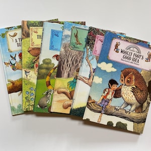 May include: A stack of vintage children's books with colorful illustrations. The covers feature titles like "Woolly Foot's Good Idea" and "Toddy and the Fox." The artwork includes images of animals, trees, and whimsical characters. The books are arranged on a white surface.