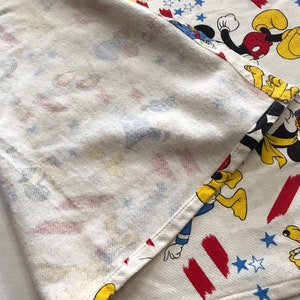 May include: White fabric with a red, white and blue pattern featuring Mickey Mouse, Donald Duck and Goofy. The fabric is folded in half, showing the back side.