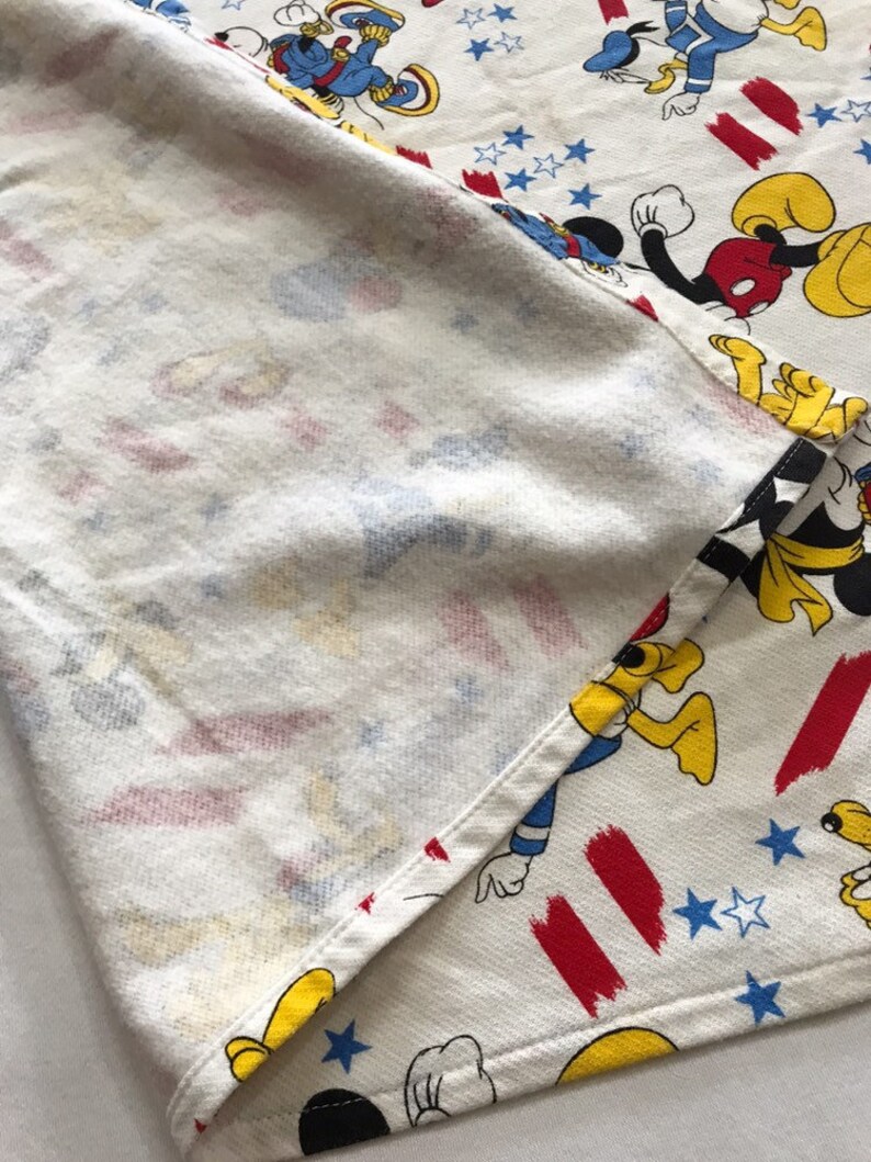 May include: White fabric with a red, white and blue pattern featuring Mickey Mouse, Donald Duck and Goofy. The fabric is folded in half, showing the back side.