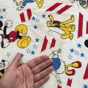 May include: White fabric with a red, white and blue pattern featuring cartoon characters such as Mickey Mouse, Donald Duck and Pluto. The fabric is covered in stars and stripes.