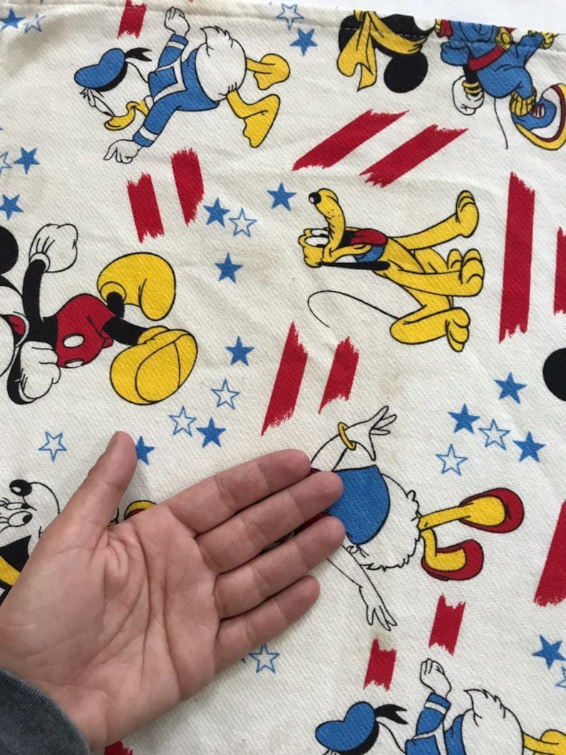 May include: White fabric with a red, white and blue pattern featuring cartoon characters such as Mickey Mouse, Donald Duck and Pluto. The fabric is covered in stars and stripes.