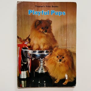 May include: A book cover with the title "Playful Pups" in blue text on a brown background. Two fluffy, orange Pomeranian dogs are pictured in front of a silver trophy. The book is part of the "Toppan's Foto Books" series.