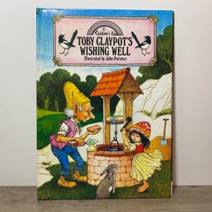 May include: A hardback children's book titled Toby Claypot's Wishing Well, illustrated by John Patience. The cover features a gnome with a tall yellow hat, a child with a mushroom hat, and a rabbit standing by a stone wishing well.
