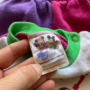 K&ouml;nnte beinhalten: Nahaufnahme eines Vintage Disney Babies Stramplers. Der Strampler hat ein Farbblock-Design mit lila, rosa, gr&uuml;nen und wei&szlig;en Abschnitten. Das Etikett zeigt Minnie- und Mickey Mouse-Illustrationen und den Text "Disney Babies".