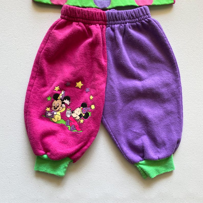 K&ouml;nnte beinhalten: Ein zweiteiliges Kinder-Outfit. Die Jogginghose ist rosa und lila mit einem Mickey- und Minnie-Mouse-Motiv und gr&uuml;nen B&uuml;ndchen. Das Oberteil ist gr&uuml;n mit rosa Punkten.
