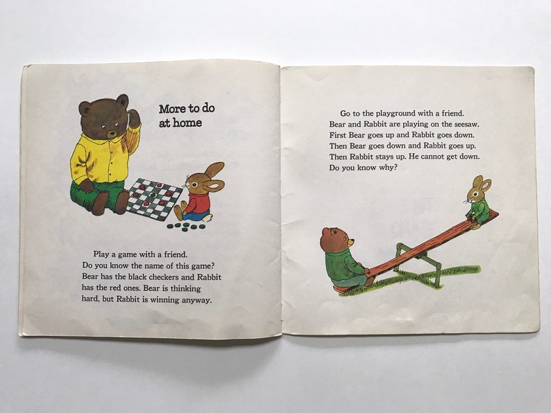May include: A children's book illustration showing a brown bear and a rabbit playing checkers and a brown bear and a rabbit playing on a seesaw. The text on the page reads "More to do at home" and "Go to the playground with a friend. Bear and Rabbit are playing on the seesaw. First Bear goes up and Rabbit goes down. Then Bear goes down and Rabbit goes up. Then Rabbit stays up. He cannot get down. Do you know why?" and "Play a game with a friend. Do you know the name of this game? Bear has the black checkers and Rabbit has the red ones. Bear is thinking hard, but Rabbit is winning anyway."