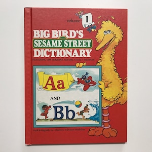 May include: A red book with a yellow Big Bird character holding a sign that says "Aa and Bb". The book is titled "Big Bird's Sesame Street Dictionary, Volume 1, Featuring Jim Henson's Sesame Street Muppets".