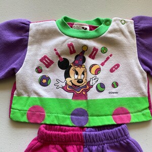 K&ouml;nnte beinhalten: Ein zweiteiliges Kinder-Outfit mit Minnie Mouse. Das Oberteil ist wei&szlig; mit einem Minnie Mouse-Motiv, dem Wort "minnie" und bunten B&auml;llen. Die &Auml;rmel sind lila und die Bord&uuml;re ist gr&uuml;n. Die Shorts sind rosa und lila.