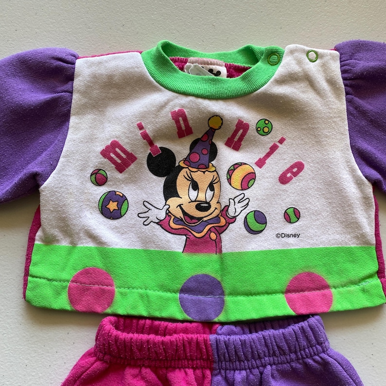 K&ouml;nnte beinhalten: Ein zweiteiliges Kinder-Outfit mit Minnie Mouse. Das Oberteil ist wei&szlig; mit einem Minnie Mouse-Motiv, dem Wort "minnie" und bunten B&auml;llen. Die &Auml;rmel sind lila und die Bord&uuml;re ist gr&uuml;n. Die Shorts sind rosa und lila.