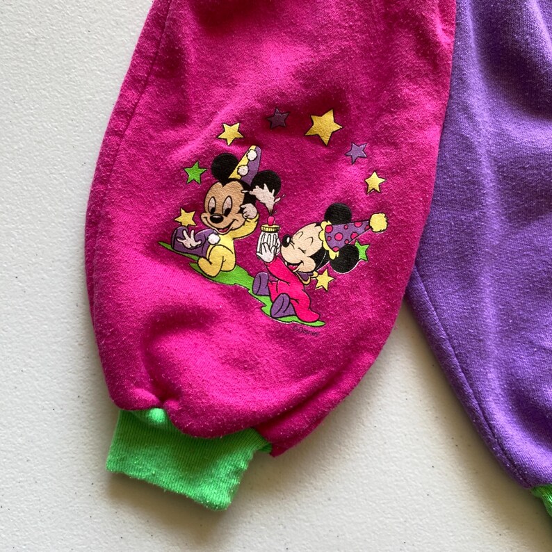 K&ouml;nnte beinhalten: Nahaufnahme eines rosa und lila Stramplers mit einer Cartoon-Grafik von Mickey und Minnie Mouse. Der rosa &Auml;rmel hat eine gr&uuml;ne Manschette und eine Grafik der Charaktere mit Sternen. Die andere Seite des Stramplers ist lila.