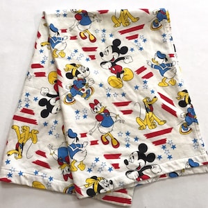 May include: A white cotton blanket with a repeating pattern of Mickey Mouse, Donald Duck, Goofy, and stars. The characters are in red, yellow, blue, and black on a white background. The blanket has red and white stripes.