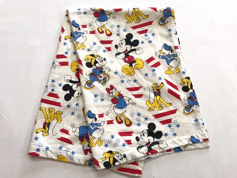 May include: A white cotton blanket with a repeating pattern of Mickey Mouse, Donald Duck, Goofy, and stars. The characters are in red, yellow, blue, and black on a white background. The blanket has red and white stripes.