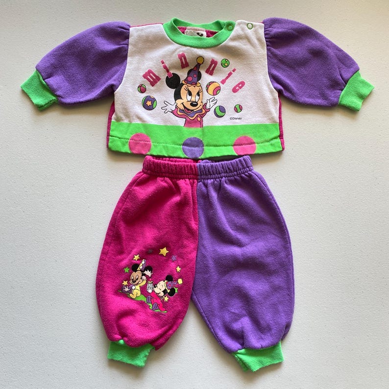 K&ouml;nnte beinhalten: Ein zweiteiliges Kinder-Outfit mit einem wei&szlig;en, lila und gr&uuml;nen Oberteil mit Minnie Mouse-Grafik und einer passenden rosa und lila Hose. Das Oberteil hat lange lila &Auml;rmel mit gr&uuml;nen B&uuml;ndchen, und die Hose hat gr&uuml;ne B&uuml;ndchen. Das Outfit ist aus einem weichen Material.