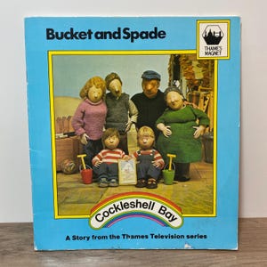 May include: A vintage children's book titled "Bucket and Spade" with a blue cover. The cover features a colorful illustration of a family of claymation figures. The book is from the Thames Television series "Cockleshell Bay."