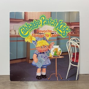 Peut inclure: Un livre Cabbage Patch Kids intitulé Favorite Things to Do! montrant une poupée aux cheveux en laine jaune, nœuds bleus et robe à fleurs bleues tenant un livre Fish World près d'un bocal à poisson rond sur un support en métal fin.