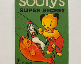 以下が含まれることがあります： 緑色の表紙の「Sooty's Super Secret」というタイトルのヴィンテージ子供向けの本。表紙には、クマのSooty、白い犬、大きなオレンジ色の魚の漫画イラストが描かれています。Sootyは釣り竿を持っています。この本はPurnellによって出版されました。
