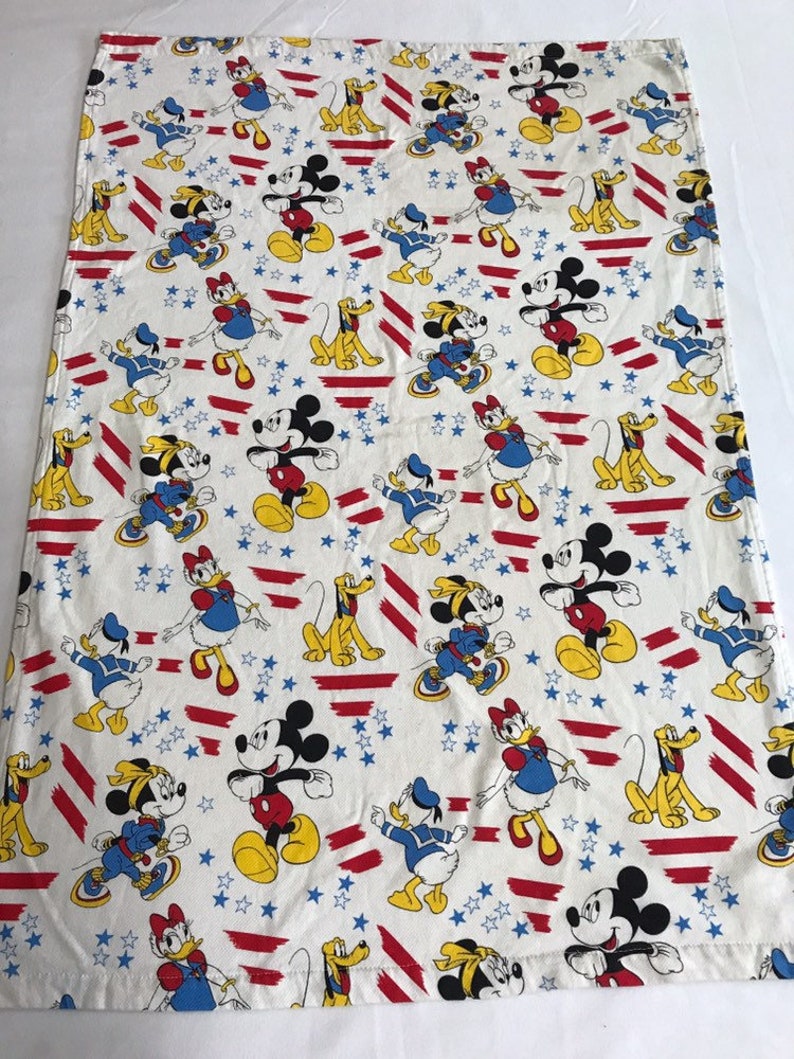 May include: A white cotton fabric with a repeating pattern of Mickey Mouse, Minnie Mouse, Donald Duck, and Pluto. The characters are wearing red, white, and blue clothing and are standing on a background of red and white stripes and blue stars.