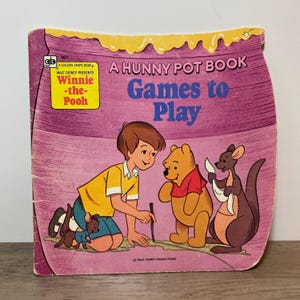 May include: A vintage children's book, "A Hunny Pot Book Games to Play", featuring Winnie-the-Pooh and friends. The cover has a purple background with illustrations of the characters. It is a Golden Shape Book.