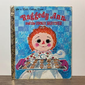 May include: A vintage Little Golden Book titled "Raggedy Ann and the Cookie Snatcher." The cover features a cartoon illustration of Raggedy Ann holding a tray of cookies. The background is blue with a checkered pattern. The book's spine is gold.