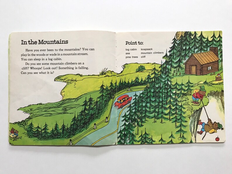 May include: An illustrated children's book page showing a red car driving on a winding road through a green forest. The text on the page reads "In the Mountains" and "Point to: log cabin, knapsack, mountain climbers, pine trees, cliff".