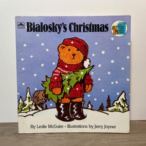 May include: A vintage children's book cover for "Bialosky's Christmas." The illustration shows a brown bear in a red hat, sweater, and boots, carrying a Christmas tree. The background is a blue snowy scene with snowflakes and pine trees.