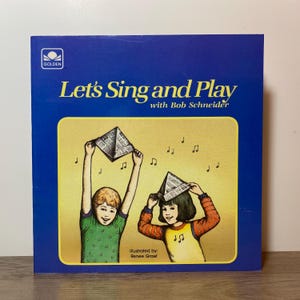 May include: A vintage children's book cover for "Let's Sing and Play with Bob Schneider." The illustration shows two children with paper hats and musical notes. The cover is blue with a yellow frame, and the Golden logo is visible.
