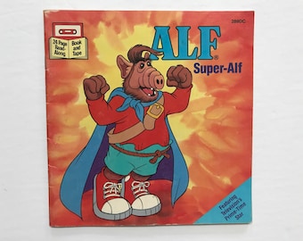 Può includere: Una copertina di libro con ALF, un personaggio alieno peloso, vestito da supereroe con un mantello rosso e pantaloni blu. Il titolo del libro è "ALF Super-Alf" ed è un libro di 24 pagine da leggere con un nastro.