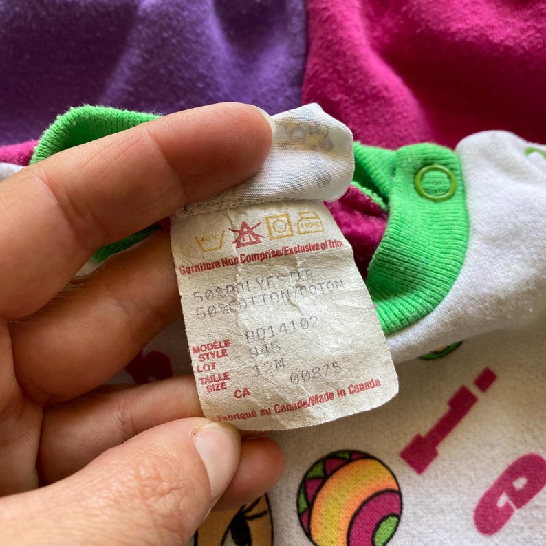 K&ouml;nnte beinhalten: Nahaufnahme eines Baby-Stramplers mit Etikett. Der Strampler hat eine wei&szlig;e Basis mit bunten Cartoon-Grafiken. Das Etikett zeigt Waschanweisungen, Stoffzusammensetzung und Gr&ouml;&szlig;eninformationen. Der Strampler hat gr&uuml;ne Verzierungen und rosa und lila Akzente.
